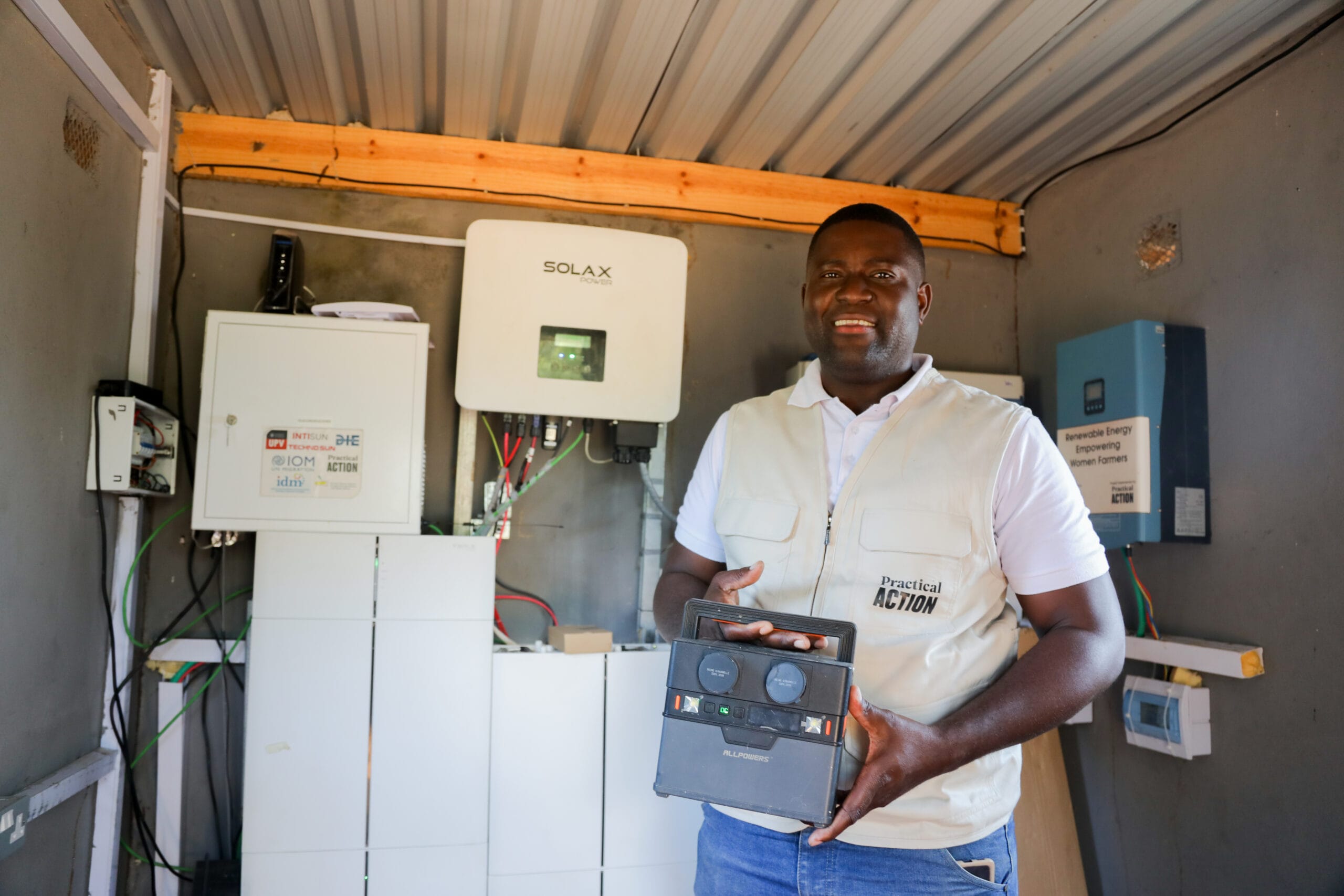 A man in a white vest stands inside a small building holding a solar device, with solar power equipment and electrical panels mounted on the wall behind him.