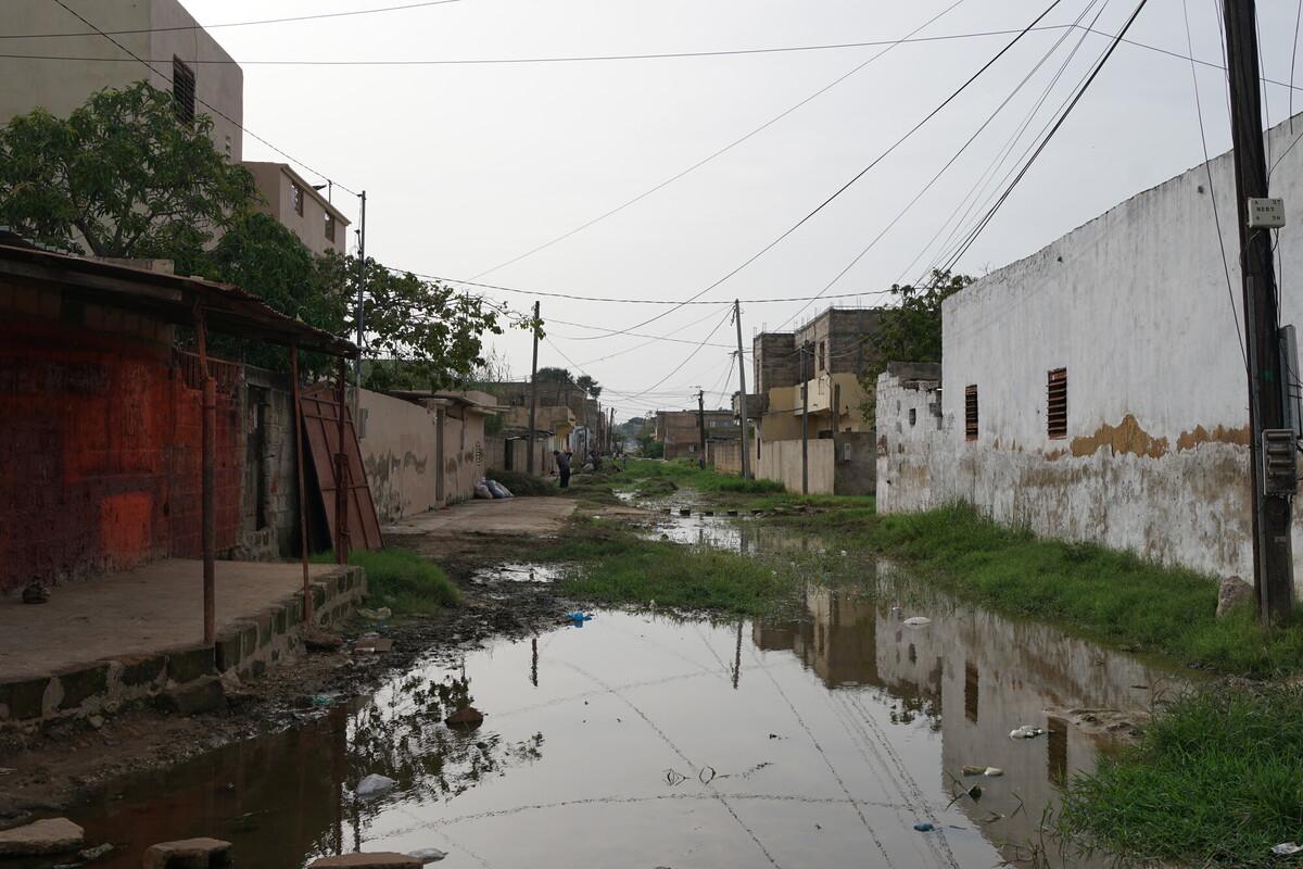 A narrow, muddy street in a residential area is flooded with water. Houses with worn walls line both sides, and grass grows along the edges. Power lines and poles are visible under an overcast sky.