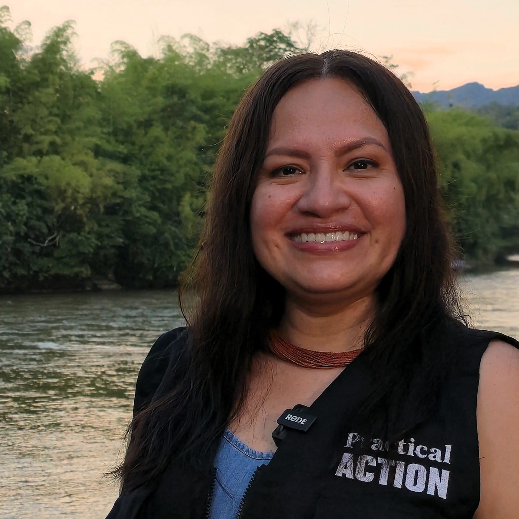 A woman with long dark hair and a sleeveless waistcoat labelled "Practical Action" smiles outdoors near a river, with green trees and mountains in the background at sunset.