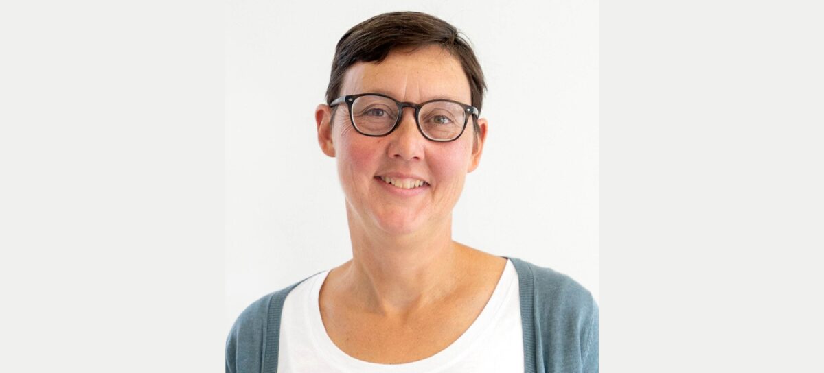 A person with short brown hair, wearing glasses, a white shirt, and a blue cardigan, is smiling in front of a plain light background.