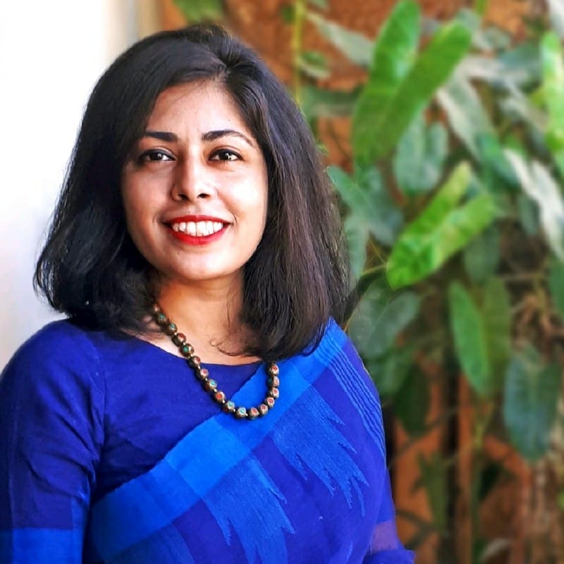 A woman with shoulder-length black hair, wearing a blue sari and a beaded necklace, smiles whilst standing indoors. Green leafy plants are visible in the blurred background.