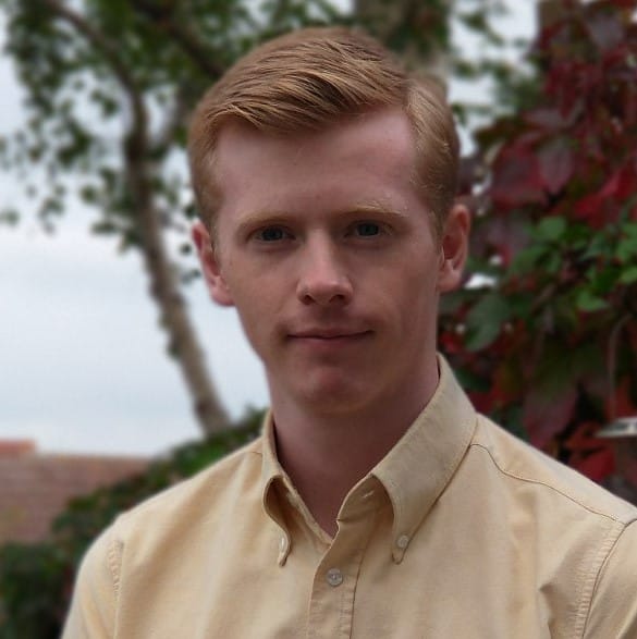 A young man with short, neatly styled ginger hair wearing a light beige button-up shirt stands outdoors in front of green foliage and a tree.