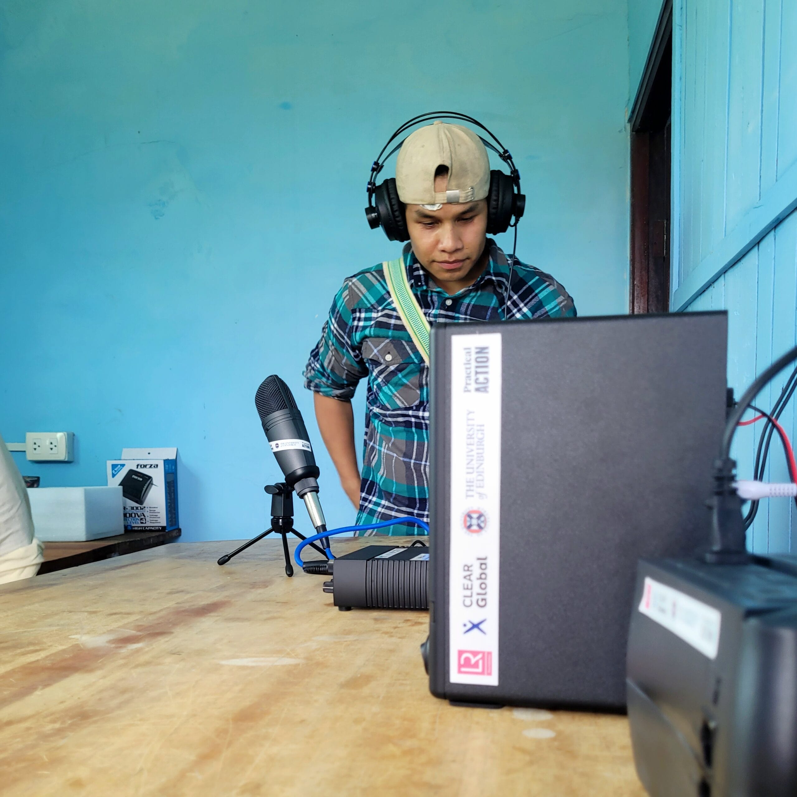A young person wearing headphones and a backwards cap stands at a wooden table with a microphone and audio equipment in a blue-walled room. Audio gear and cables are visible on the table.