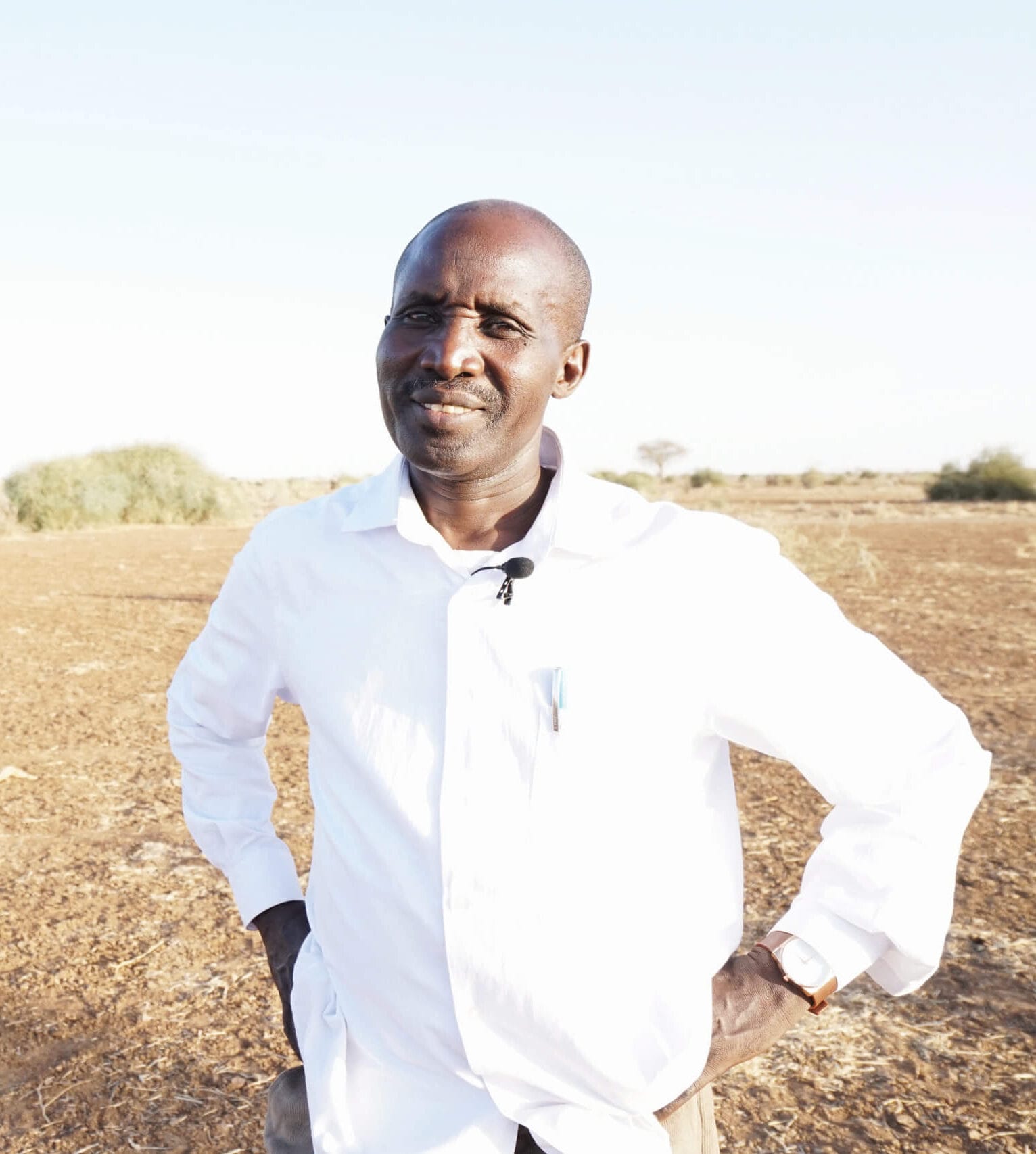 A man in a white shirt stands outdoors on dry, brown land with sparse green bushes in the background under a clear, bright sky.