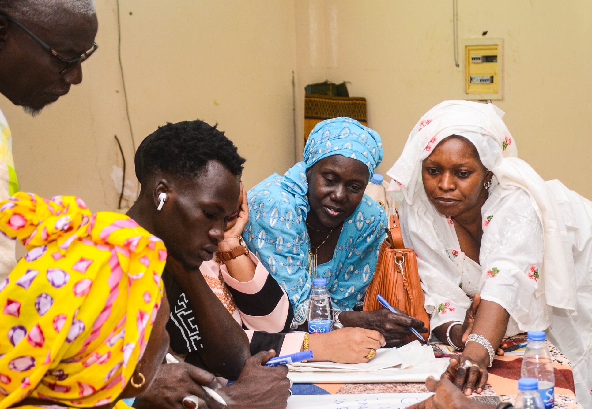 Several people, some in colourful traditional clothing, gather around a table covered with papers, pens, and water bottles, engaged in discussion and writing together indoors.