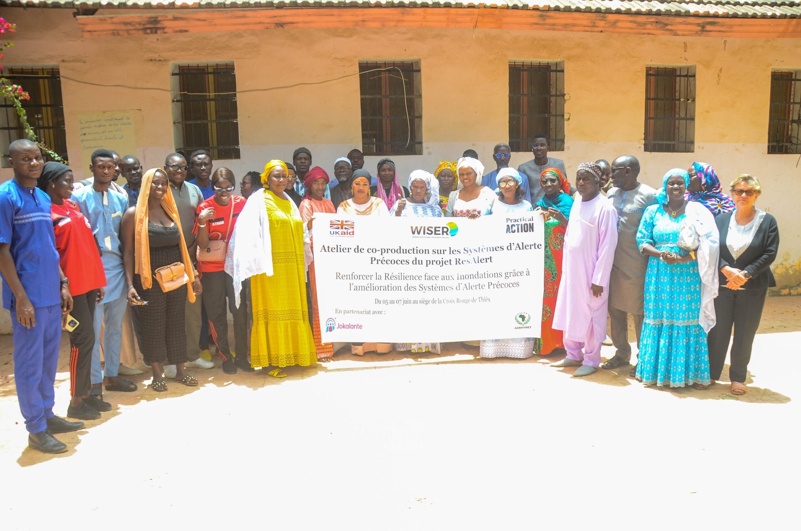 A diverse group of people stand outside a building, holding a large banner in French about reinforcing resilience and innovation in early warning systems as part of the WISER project.