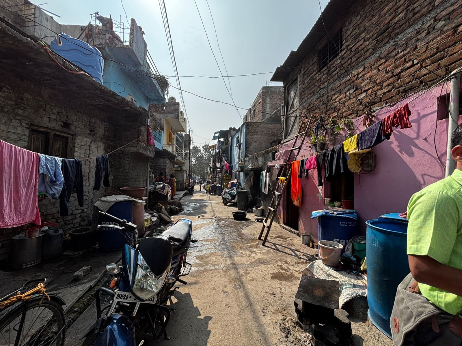 A narrow alleyway in a neighbourhood with clothes hanging on lines, colourful buildings, a parked motorbike, and various containers and items along the sides. Sunlight casts shadows on the scene.