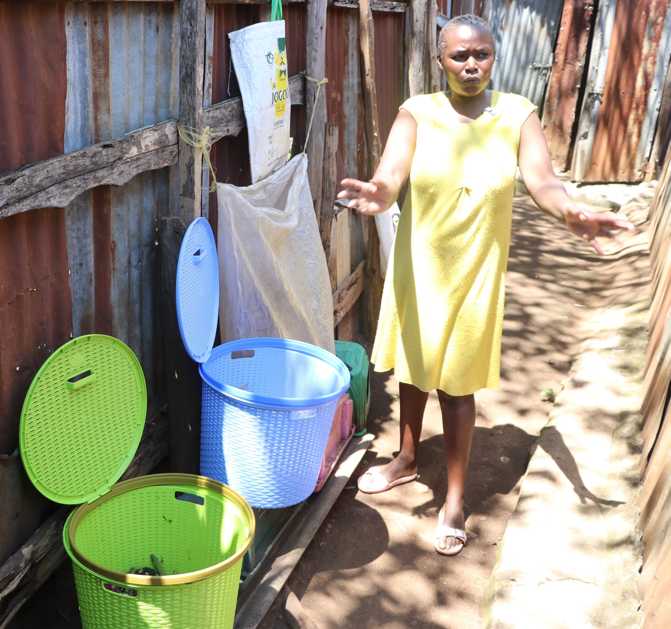 A woman in a yellow dress stands outside beside three plastic bins with open lids near a corrugated metal fence, gesturing with her arms outstretched.
