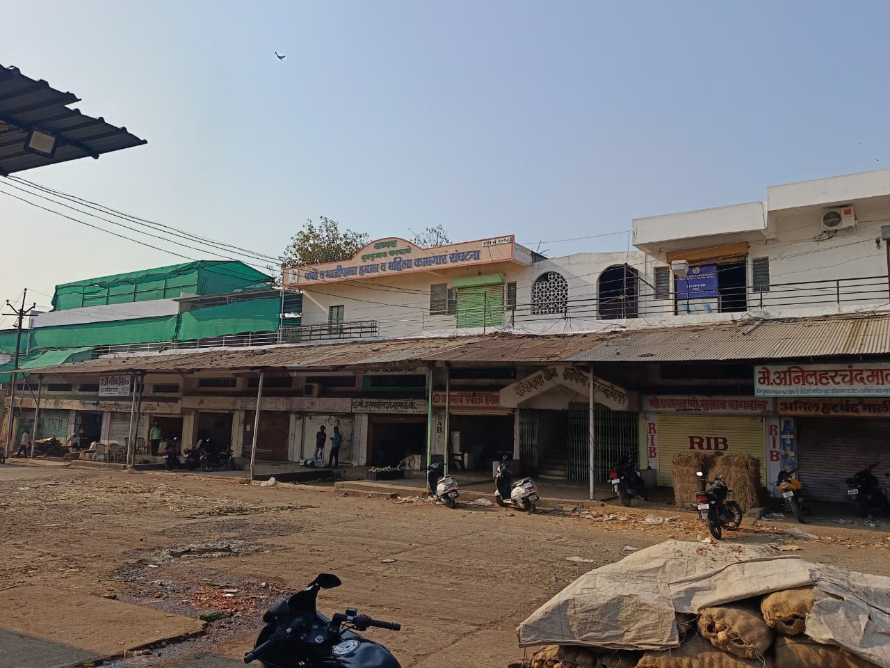 A row of shops with closed shutters, scooters parked outside, and a two-storey white building above. The area appears quiet with a few people in the shade and an empty, dusty street in front.