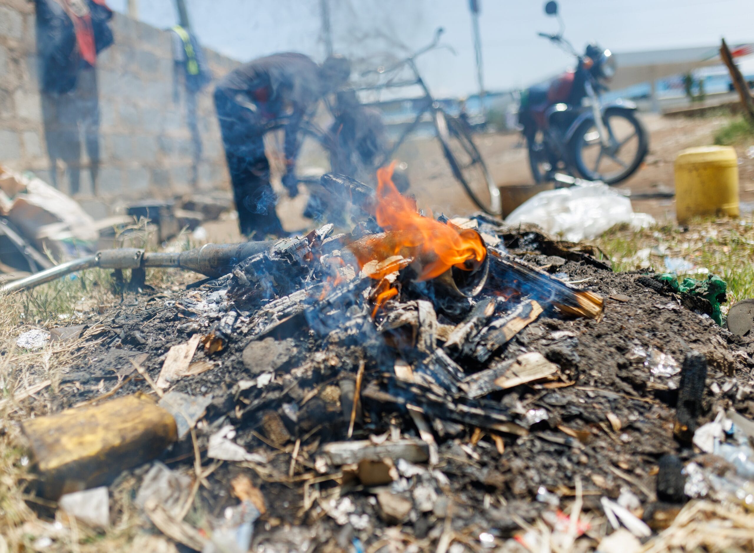 A small open fire burns rubbish and debris outdoors, with smoke rising. Blurry figures, a bicycle, and a yellow container are visible in the background, suggesting an urban or roadside setting.