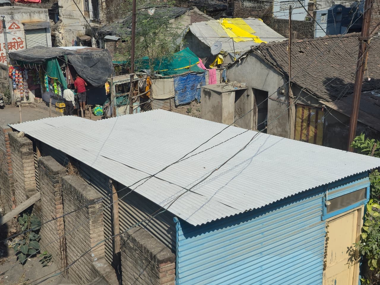 A corrugated metal roof covers brick-walled structures, with small, colourful homes and laundry hanging in the background. Power lines cross above the roof, and sunlight casts shadows on the scene.