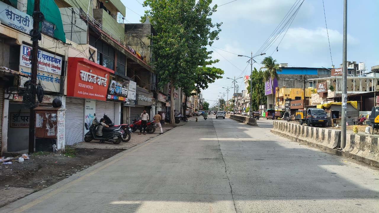A wide street in an urban area with shops, parked scooters, and a few people. Trees line the pavement, and vehicles are visible on the road. The sky is partly cloudy.