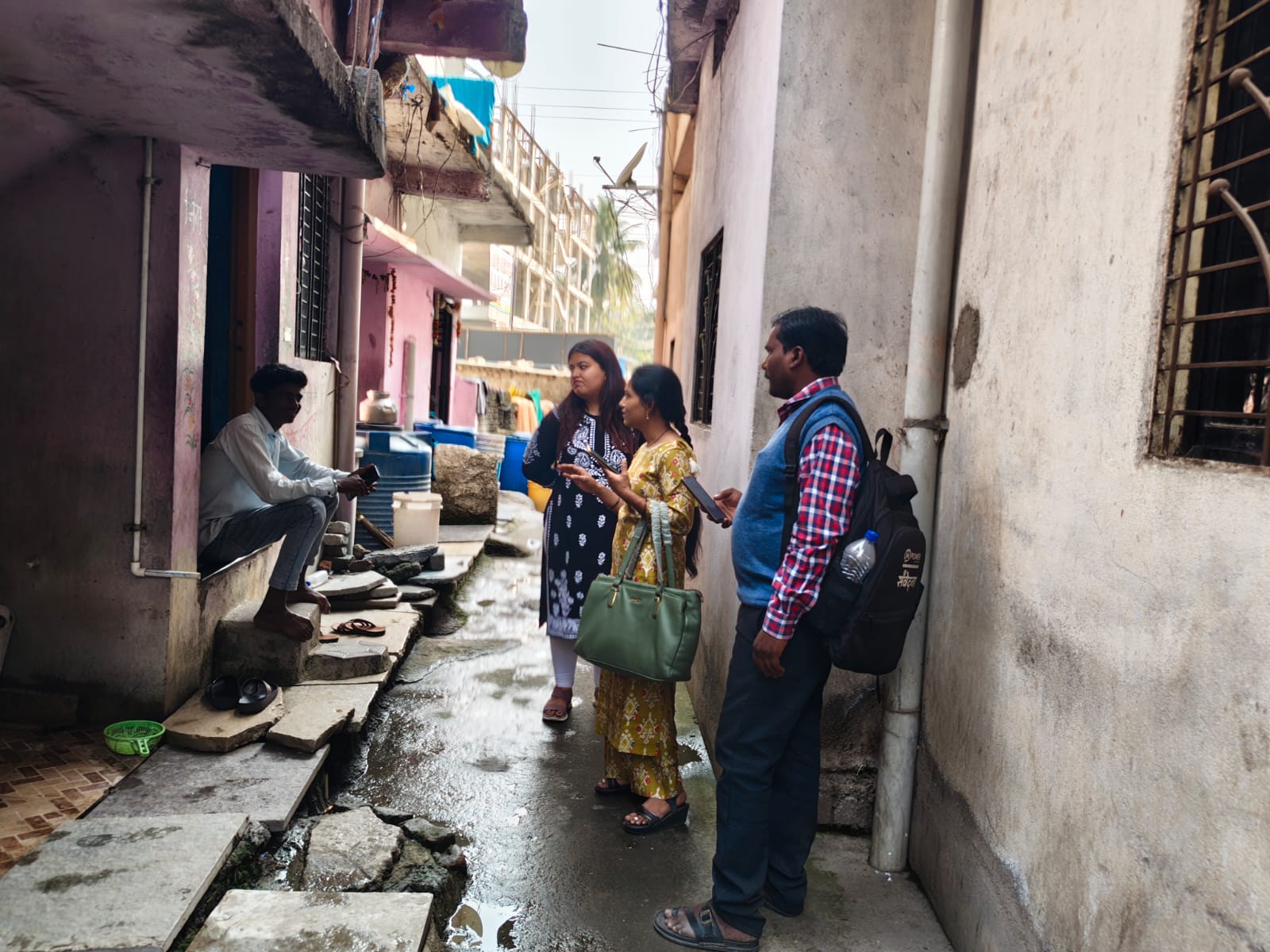 Three people stand talking to a man sitting in a narrow alleyway with uneven pavement and colourful buildings in the background.