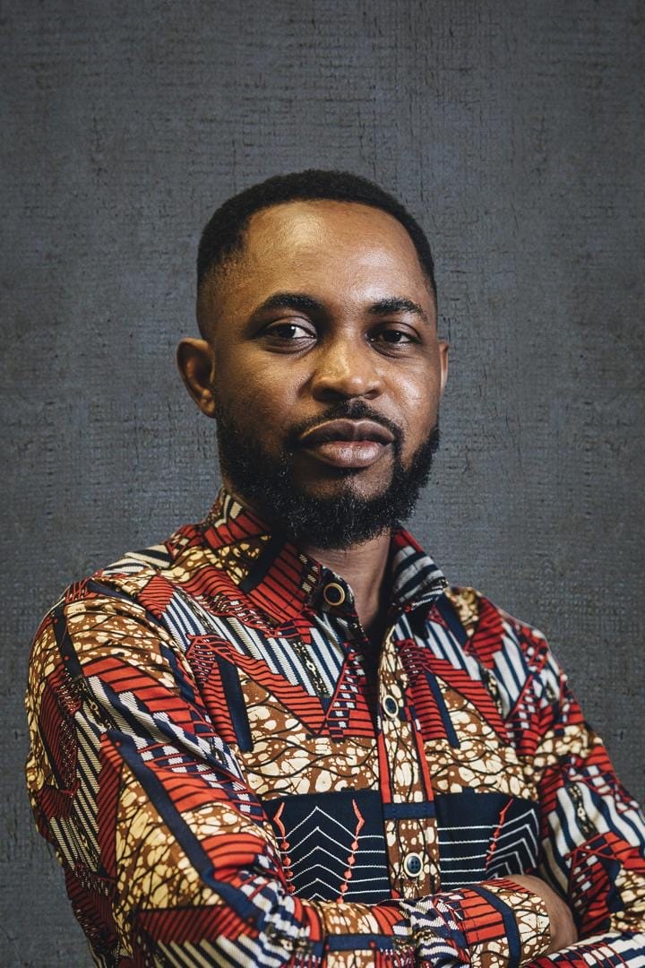 A man with a beard and moustache wearing a colourful, patterned shirt stands against a textured grey background, looking confidently slightly to the side with his arms crossed.