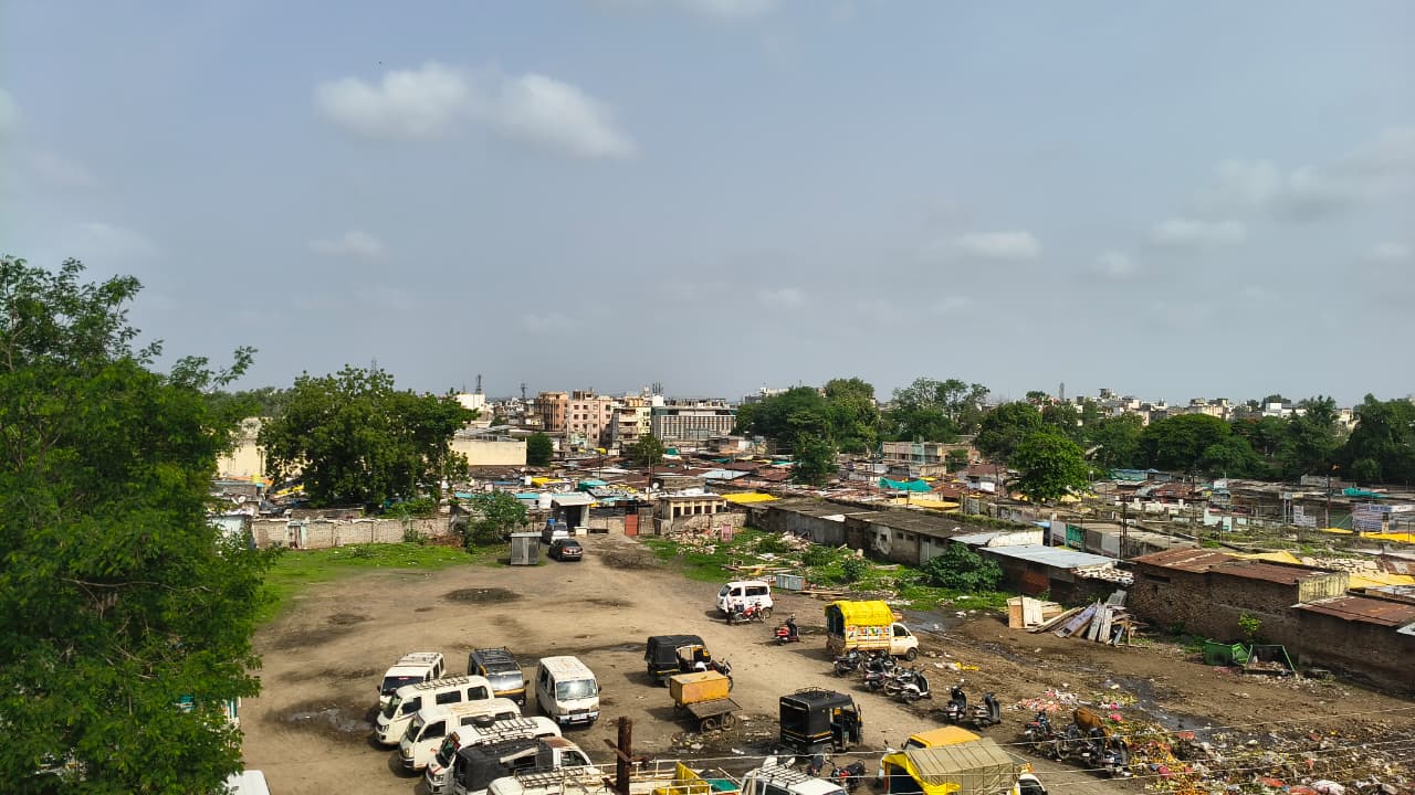 A semi-urban area with parked vehicles, scattered rubbish, small buildings, some greenery, and a blue sky with scattered clouds in the background.