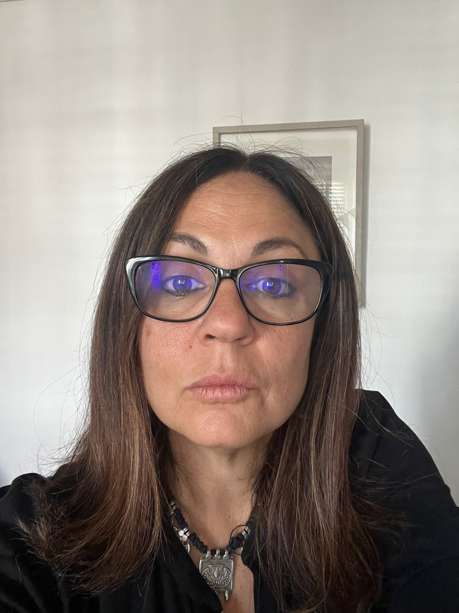 A woman with long brown hair and glasses looks at the camera. She is wearing a black top and a necklace, with a white wall and framed picture in the background.
