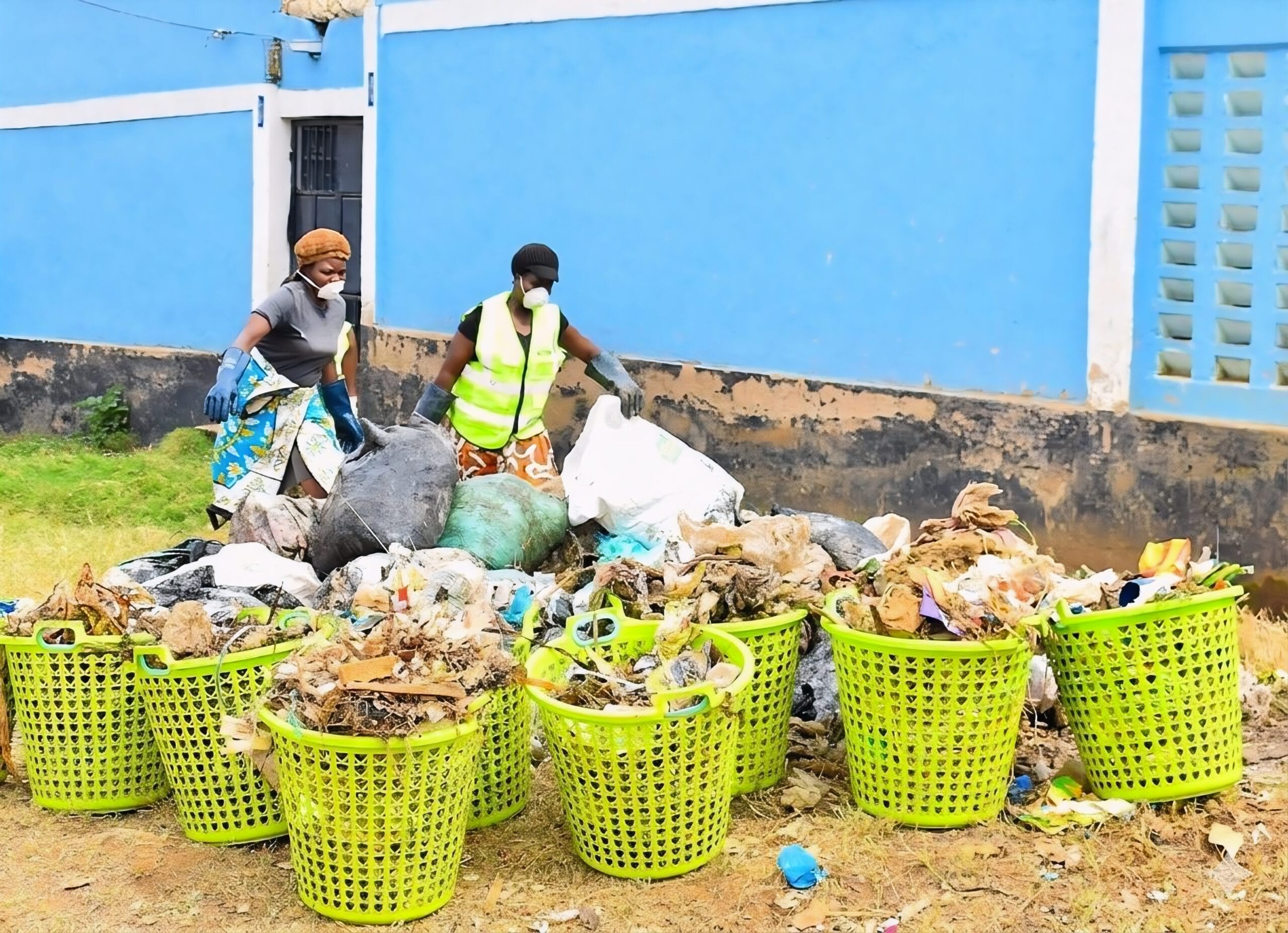 Two people wearing masks and gloves sort through piles of rubbish, placing waste into large green baskets, in front of a blue building with black trim.