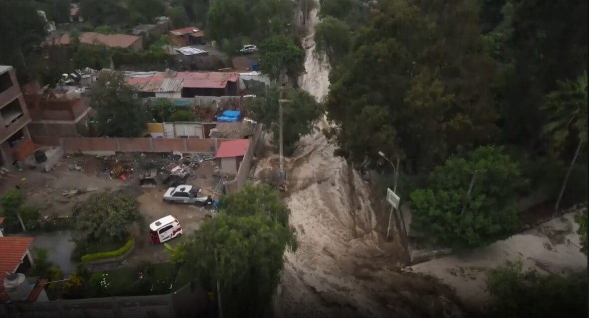 Aerial view of a neighbourhood affected by a mudslide, with muddy water flowing down a street, damaged homes and vehicles visible, and trees surrounding the area.