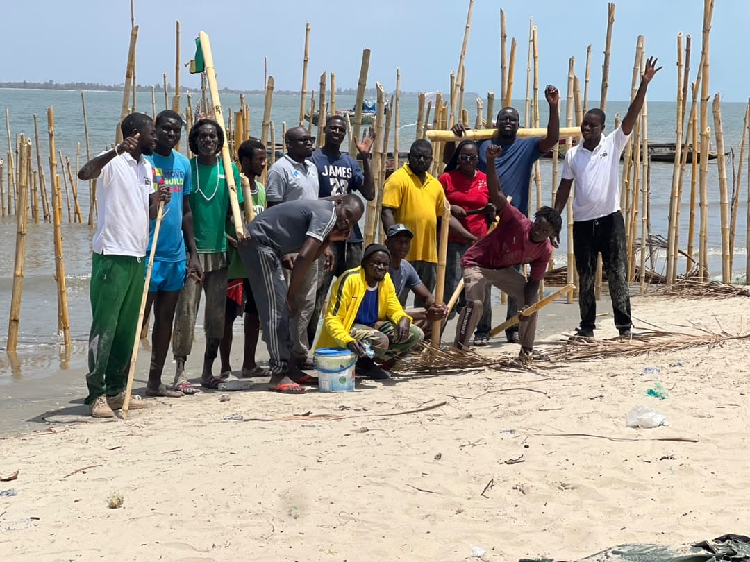 A group of people stand and crouch on a sandy beach near the water, surrounded by tall bamboo poles. Some hold poles, and a few are cheering or raising their arms. The sky is blue and it appears sunny.