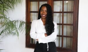 Woman in white blouse and black trousers stands smiling with arms crossed in front of window.