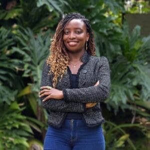 Woman with plaited hair stands smiling outside, arms folded, wearing a blazer and jeans, with green plants behind.