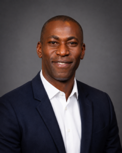 Man in navy blazer and white shirt smiling in front of a plain grey background.