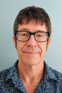 Man with short brown hair, black glasses, and a blue patterned shirt, standing against a light background.