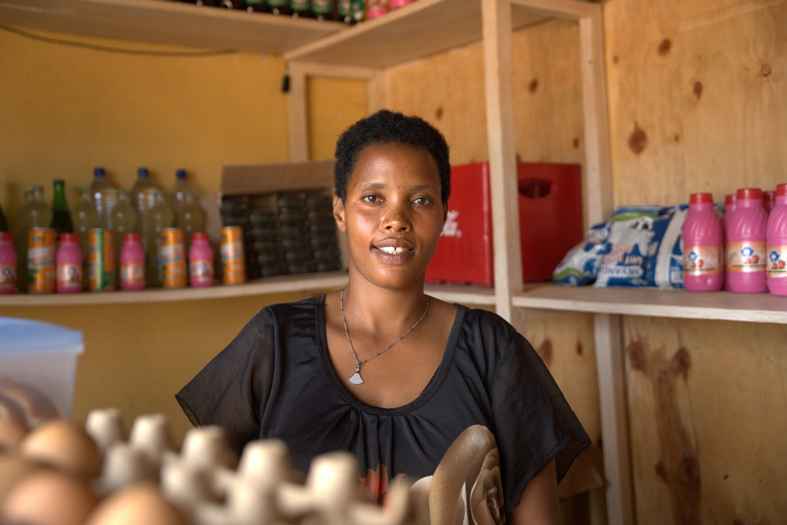Smiling woman stands in a small shop with shelves of drinks, eggs, and groceries behind her.