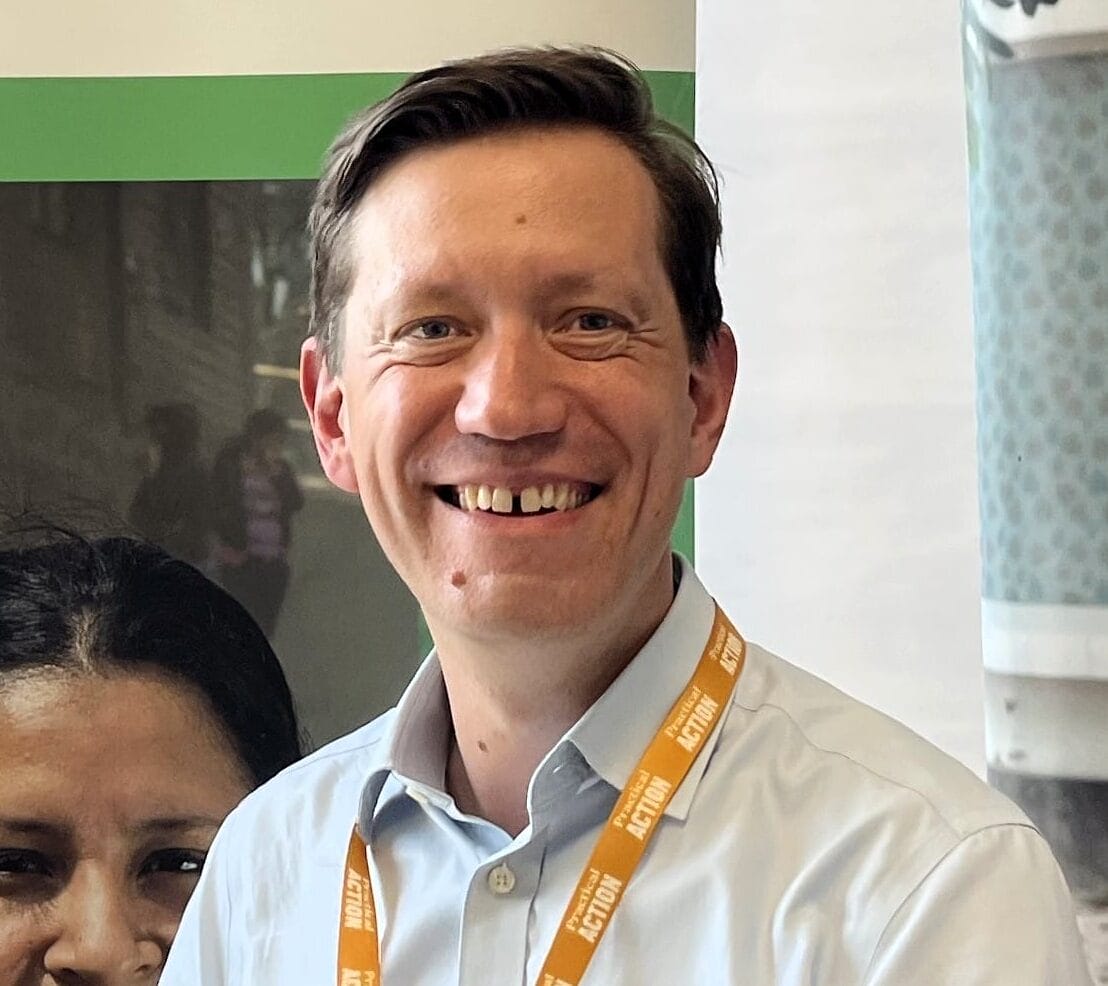 A smiling man wearing a light blue shirt and orange lanyard stands indoors in front of posters showing a woman with a spade and a person pushing a trolley.