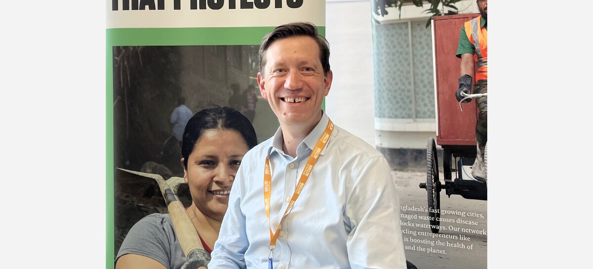 Smiling man with an orange lanyard sits in front of a project poster with images of workers.