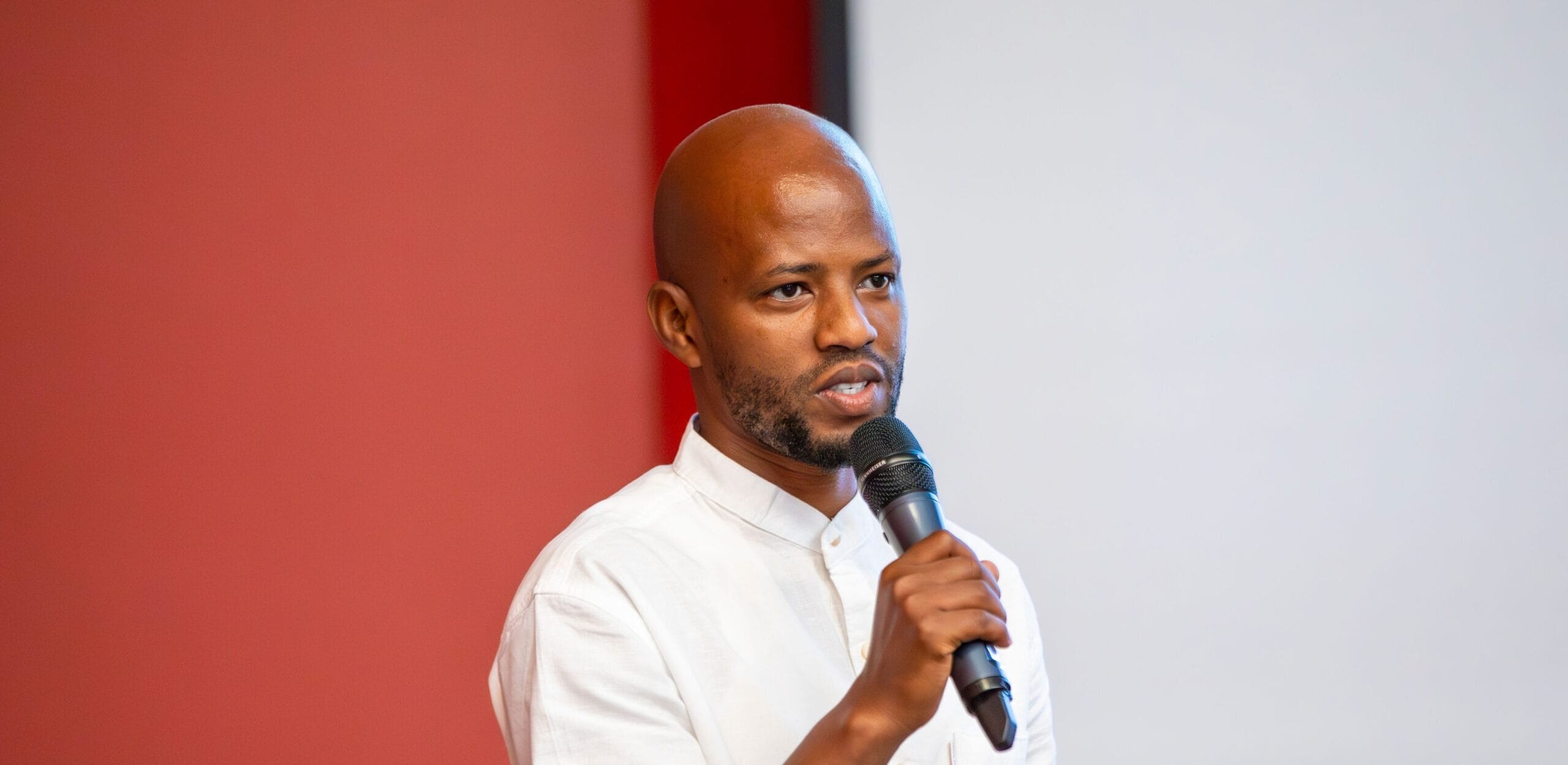 A man in a white shirt speaks into a microphone whilst standing in front of a red wall and a projection screen.