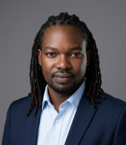 Man with long dreadlocks wearing a dark blue suit and light blue shirt, posing against a grey background.