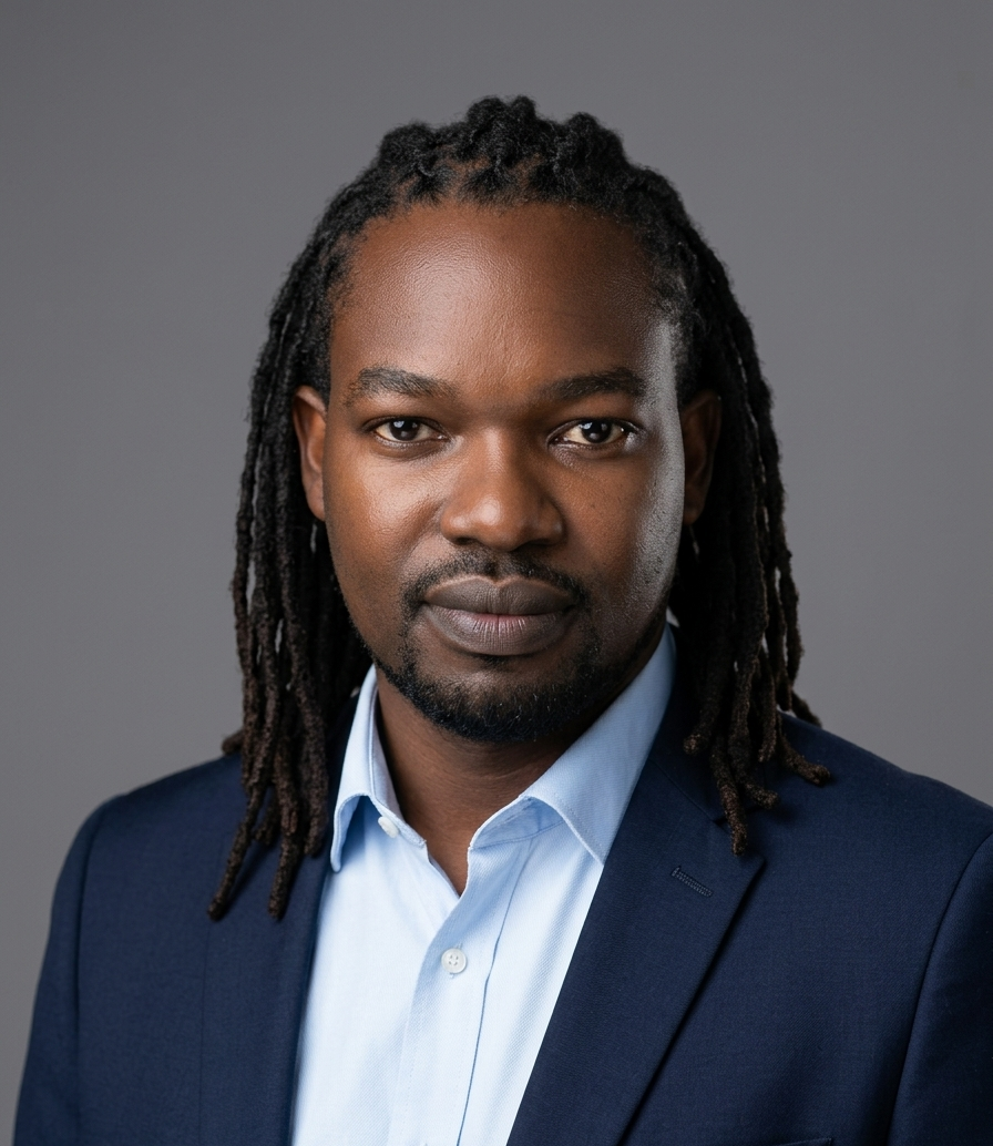 Man with long dreadlocks wearing a dark blue suit and light blue shirt, posing against a grey background.