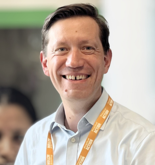 Smiling man in a light shirt wearing an orange lanyard, standing indoors with a blurred background.