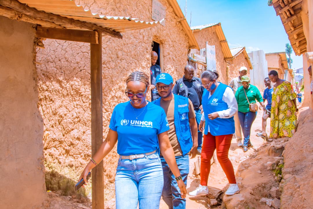 UNHCR staff walk through a sunlit village with mud houses, engaging with local residents.