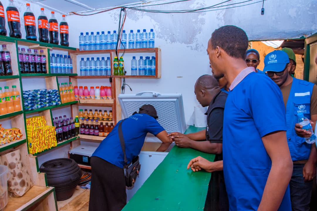 People in a small shop with drinks on shelves; one person is looking inside a freezer, others are watching.