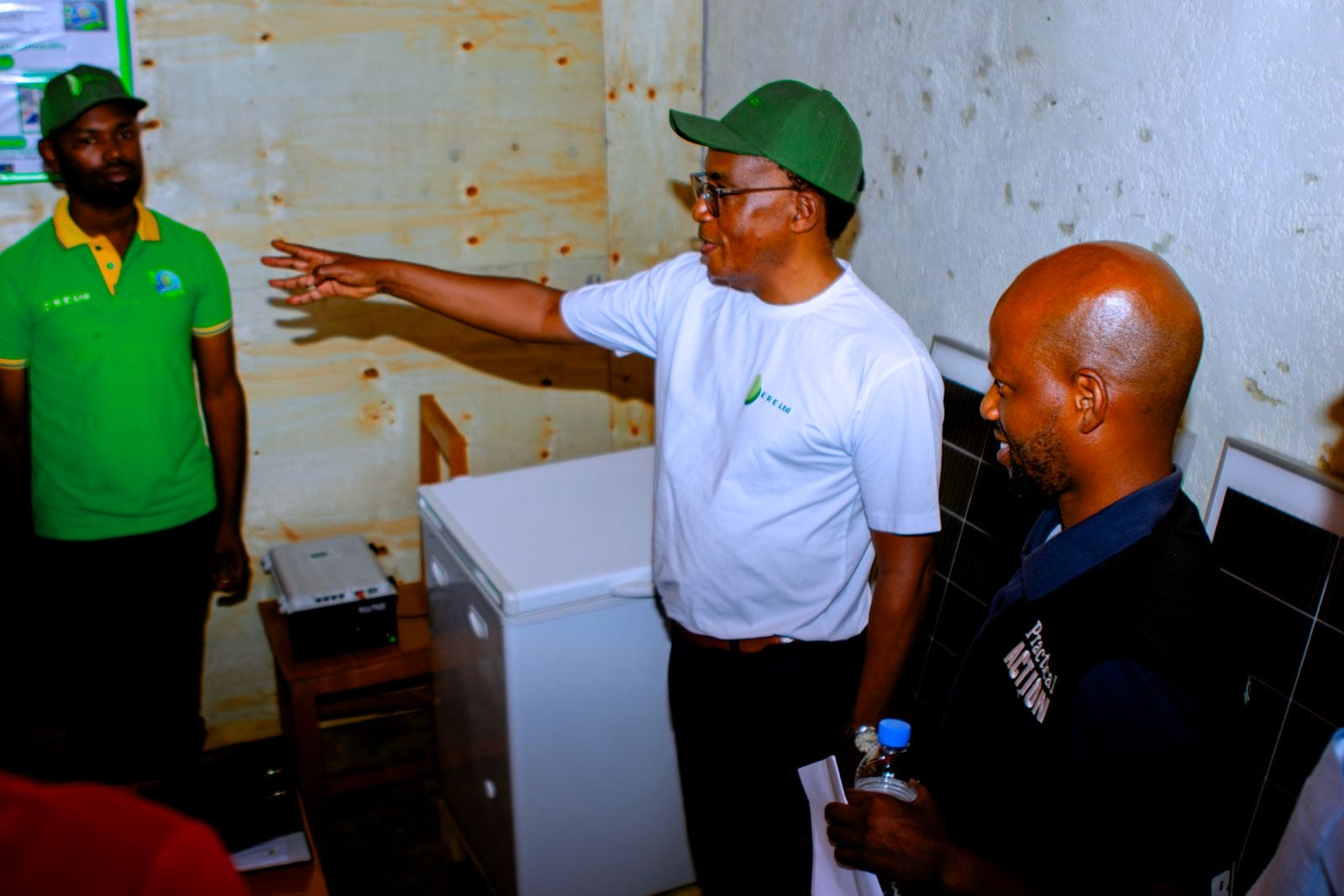 A man in a white shirt gestures whilst speaking to two others in a small room with a fridge and solar panels.