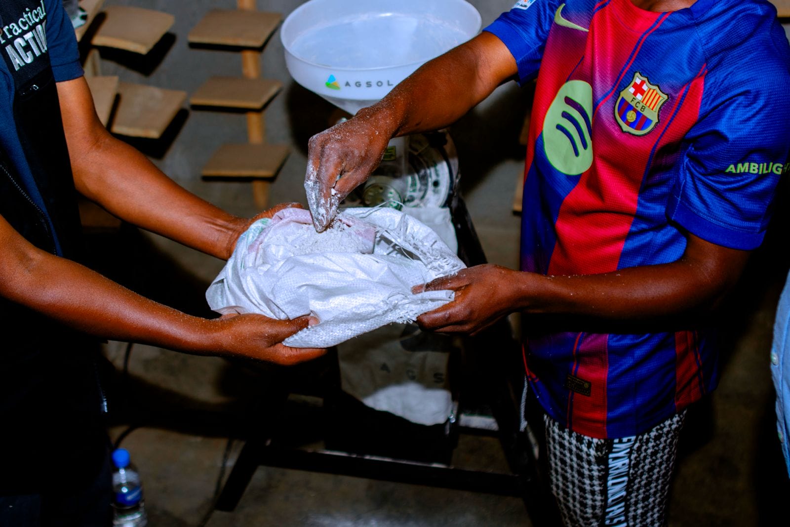 Two people pour white powder into a sack, standing near a set of scales and wearing short-sleeved shirts.