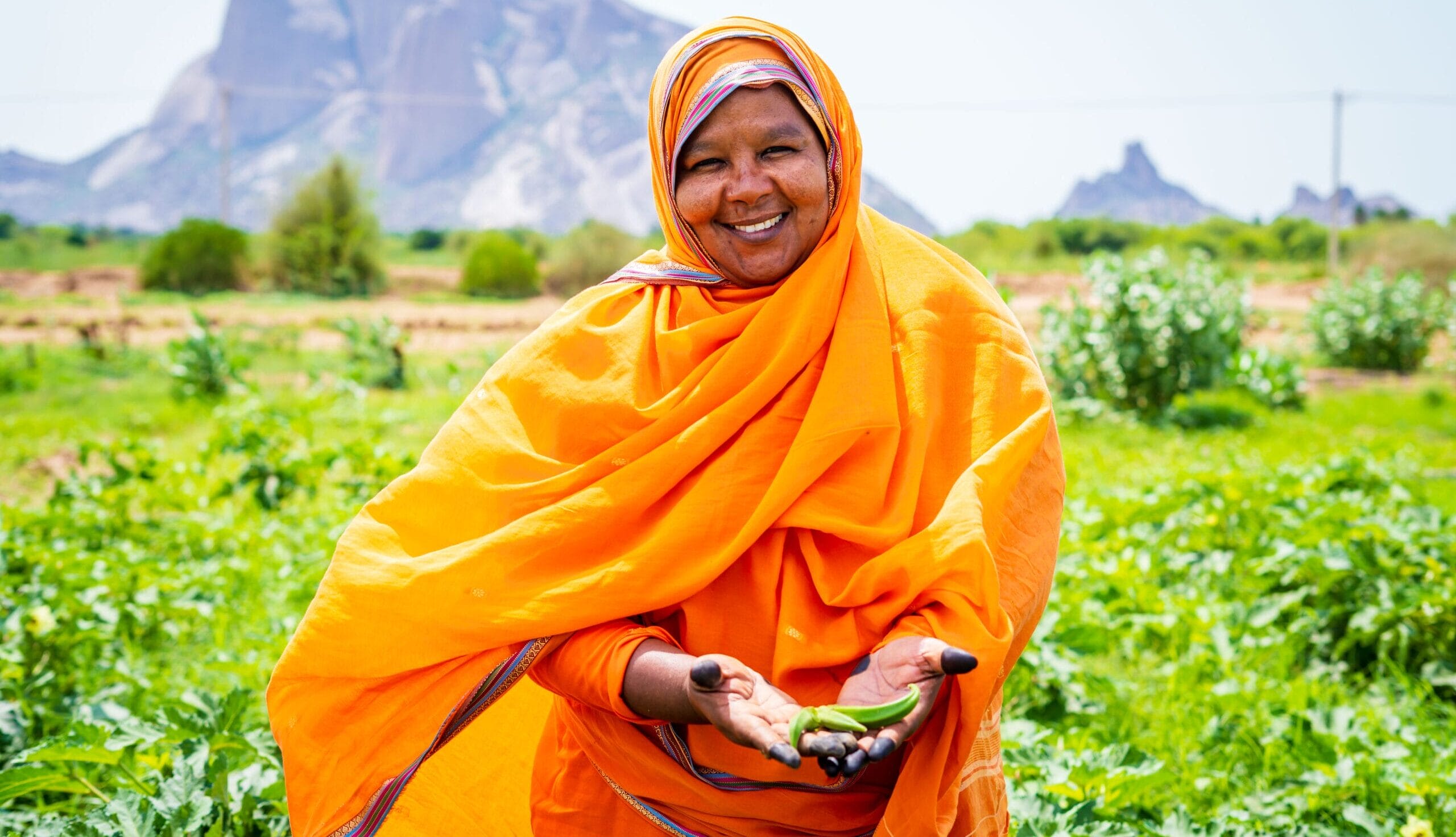 Smiling woman in an orange dress holds soil in her hands, standing in a green field with mountains behind her.