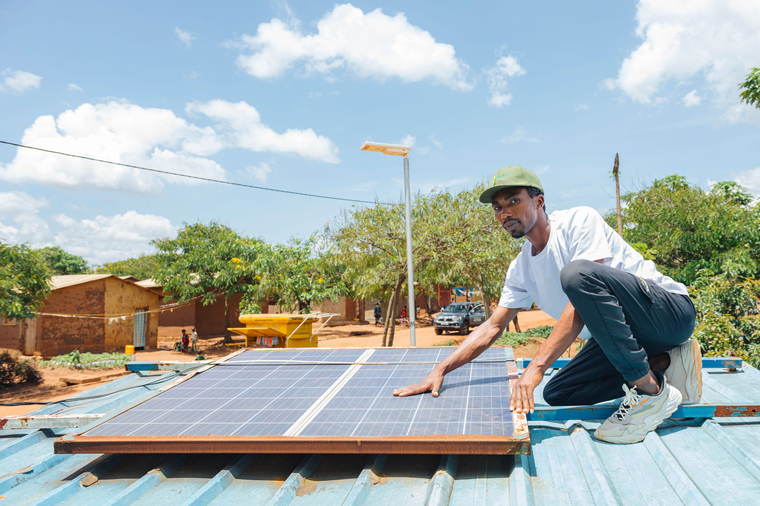 Man in a white shirt installs solar panels on a roof in a sunny rural area with trees and houses nearby.