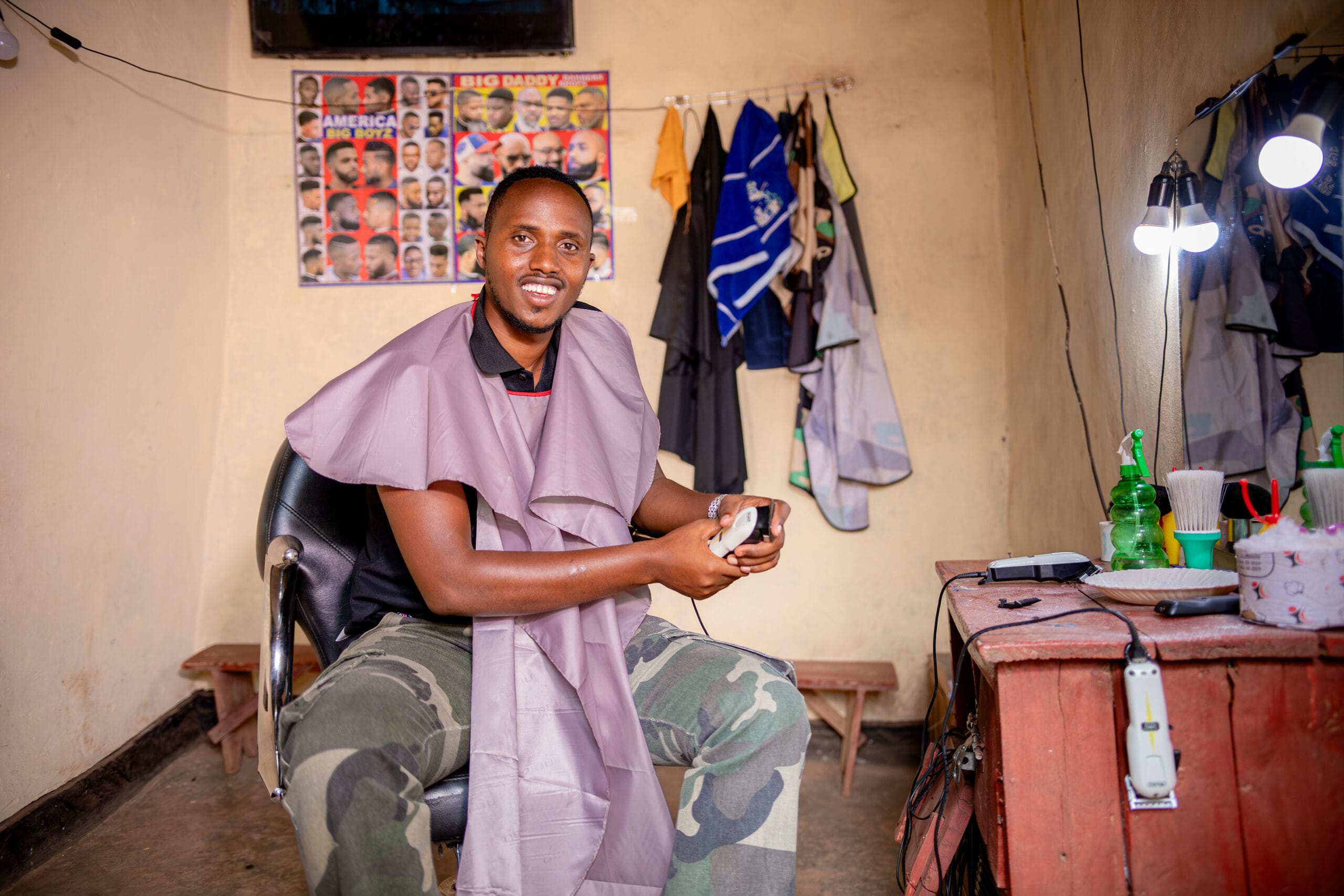 Smiling man in a barber’s shop wearing a gown, holding clippers, with barber tools on a counter nearby.