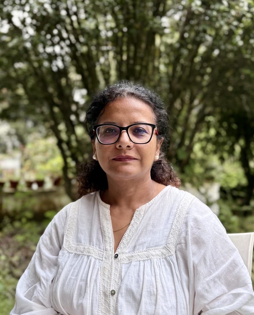 Woman with curly hair and glasses in a white blouse sits outdoors with green trees in the background.