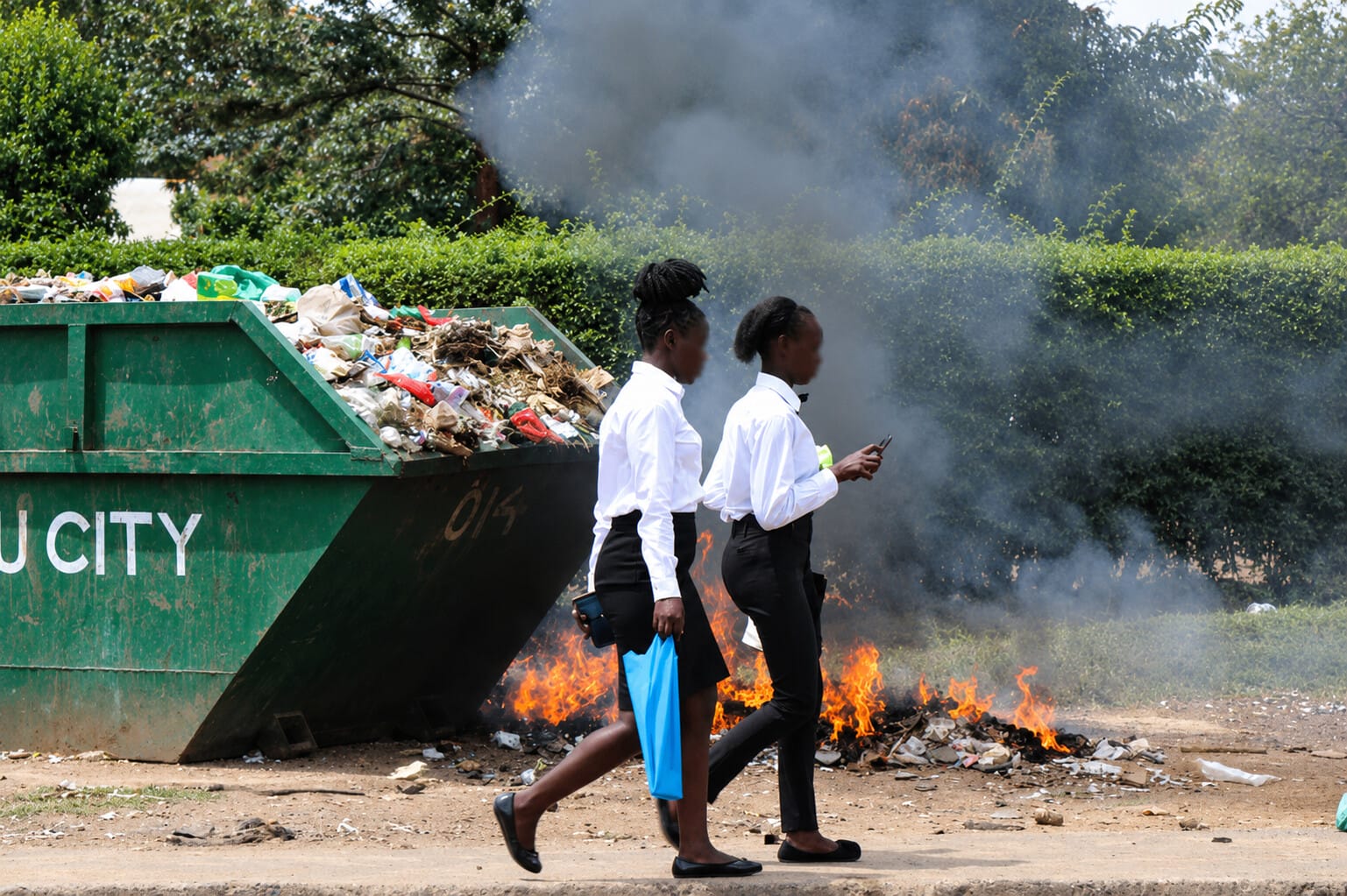 Two women walk past a burning pile of rubbish next to an overflowing skip on a city street.