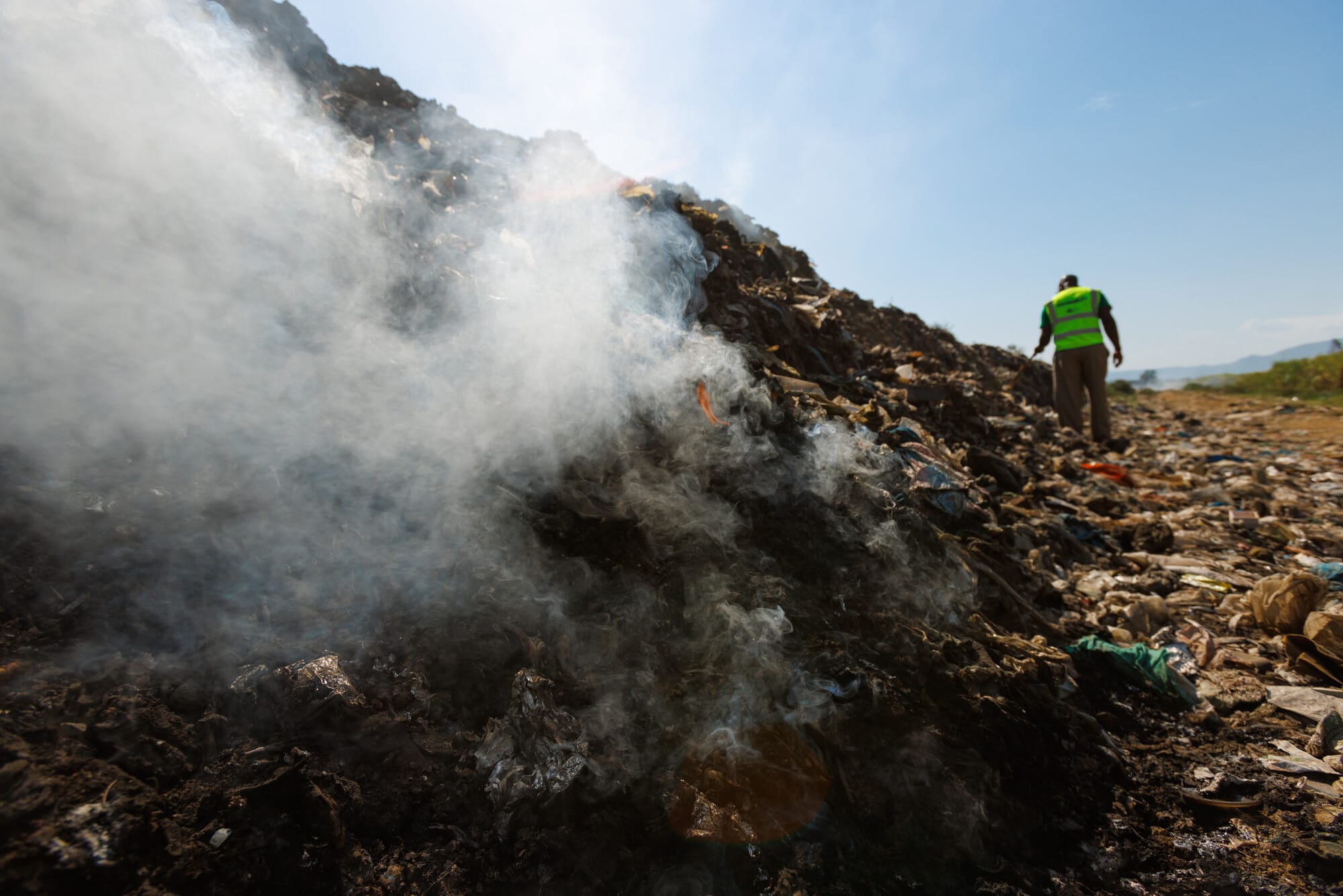 Smoke rises from a pile of burnt rubbish as a person in a green vest stands nearby in the background.