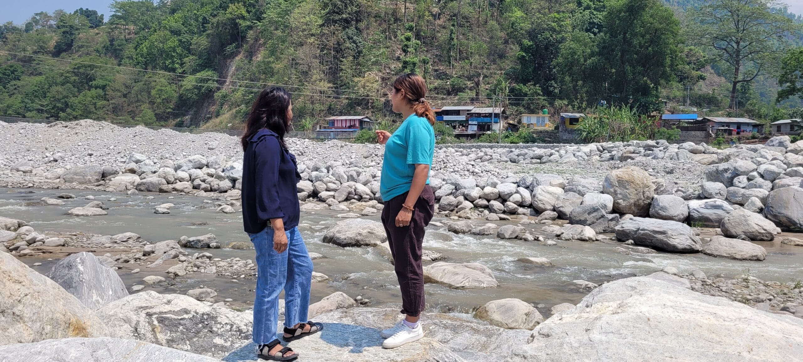 Two people stand on rocks by a river, facing each other, with trees and houses on a hillside in the background.
