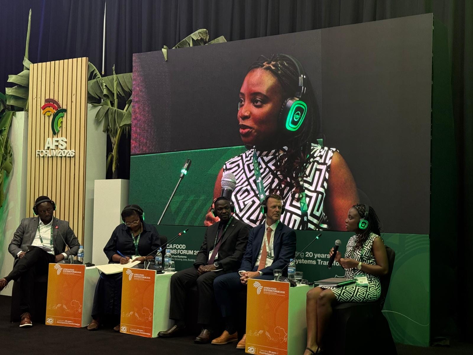 Five panellists sit on stage with a large screen showing a woman speaking, all wearing green-rimmed headsets.