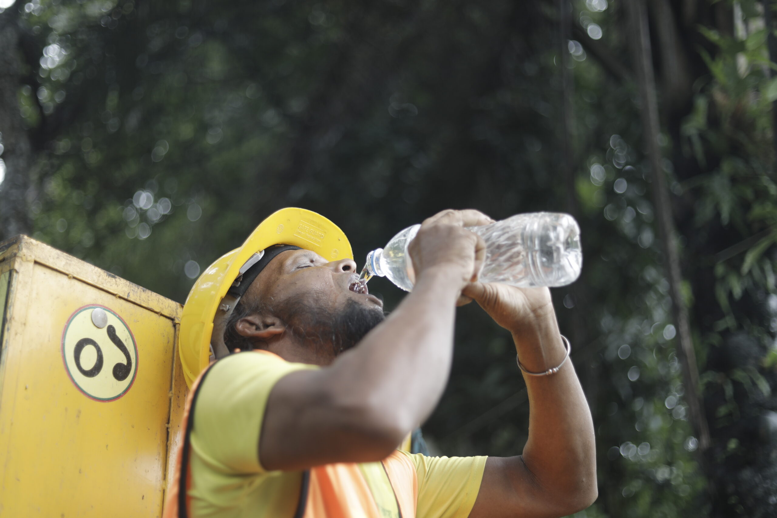 A construction worker in a yellow hard hat and safety vest drinking from a plastic water bottle.
