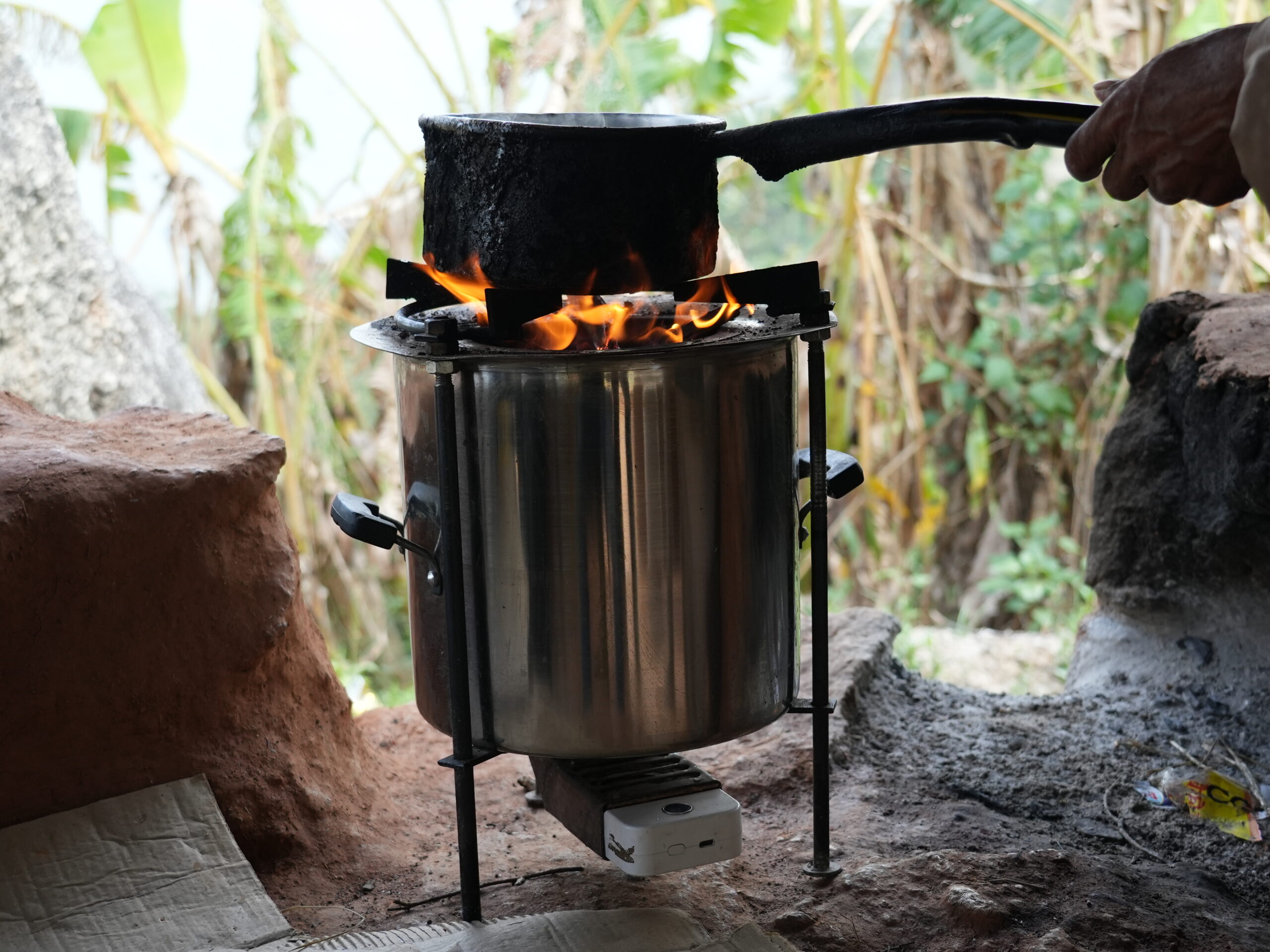 A hand holds a blackened saucepan over a metal cooker fuelled by a small fire.