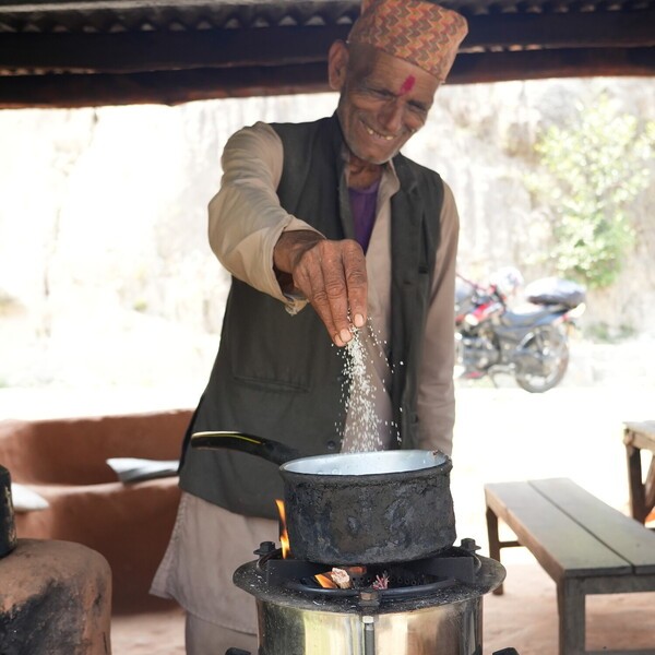 An elderly man in a traditional cap sprinkles grain into a pot over a small outdoor cooker.