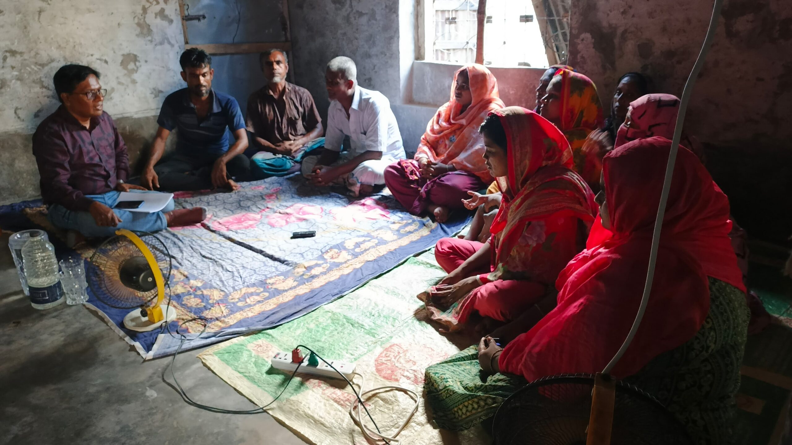 A group of people seated on floor mats in a room, listening to a man speaking.